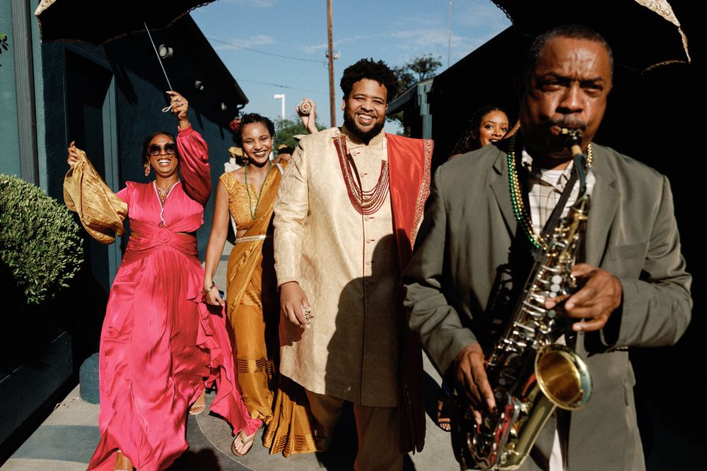 Wedding baraat procession with saxophonist leading guests in colorful Indian attire.
