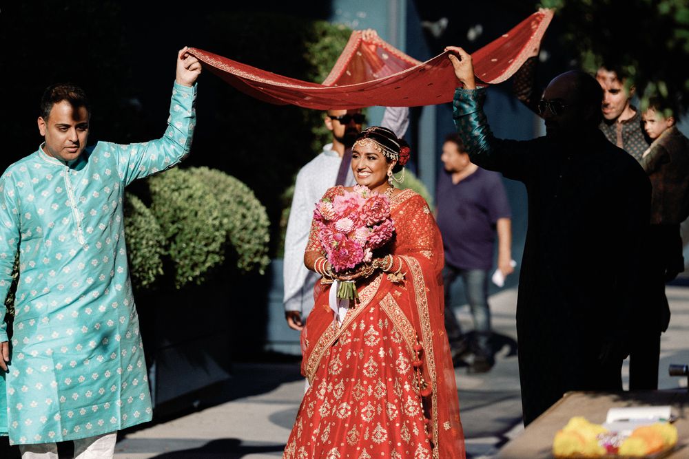 Indian bride in red lehenga walking under dupatta canopy during baraat procession.