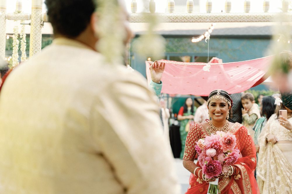 Smiling Indian bride in red lehenga walking down aisle under pink dupatta.