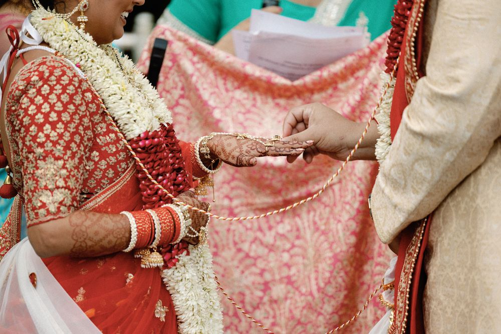 Indian bride and groom exchange hands during traditional Hindu wedding ceremony with red fabric backdrop.