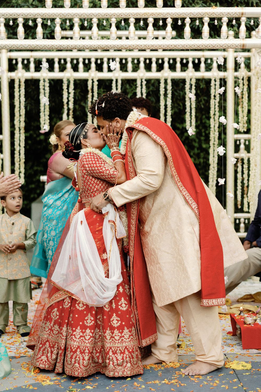 Indian bride and groom share first kiss in red and gold traditional wedding attire at outdoor ceremony.