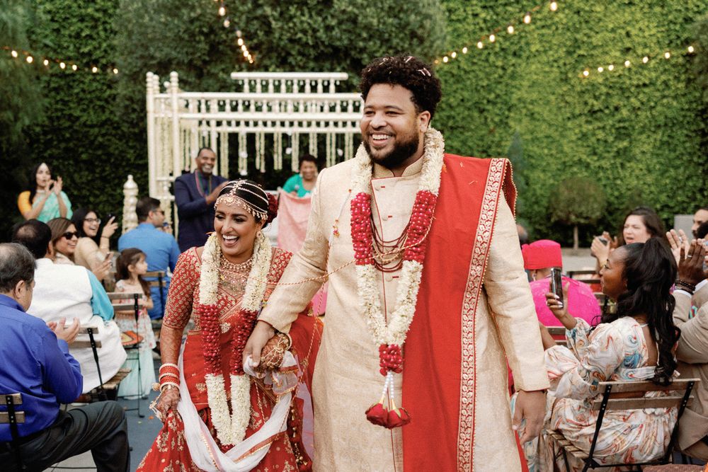 Smiling Indian newlyweds walk together in traditional red and gold wedding attire with floral garlands.