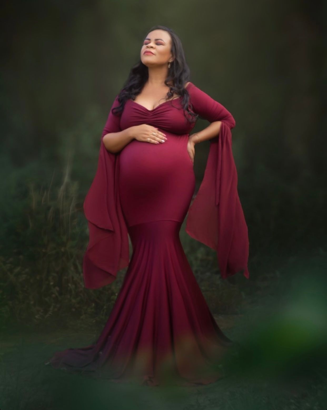 Woman in flowing burgundy dress posing gracefully against dark background.