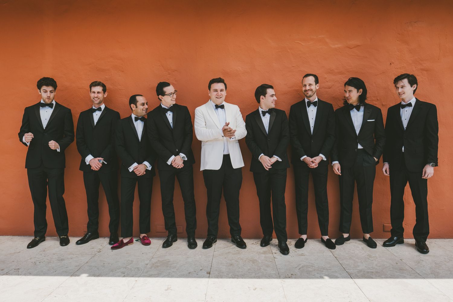 Groomsmen in white dinner jackets stand together at an elegant wedding reception with hanging greenery decorations.