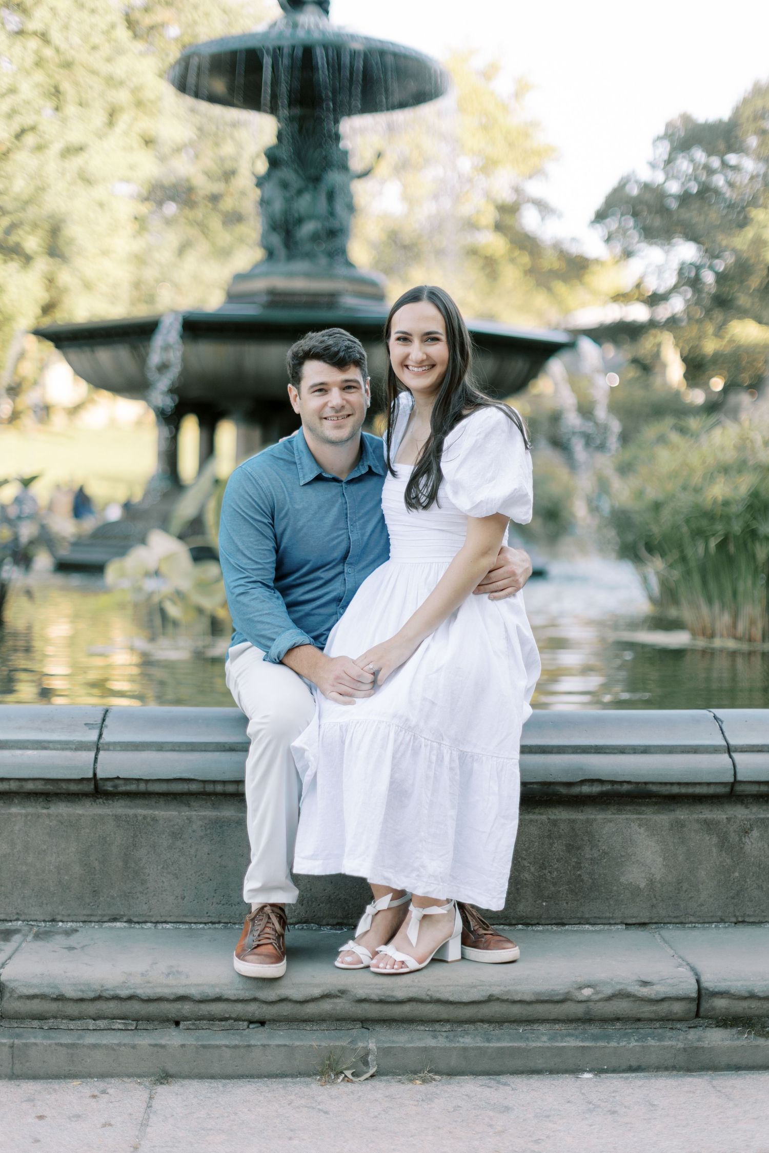 Couple sitting at edge of stone wall by pond wearing white dress and blue shirt, showing their feet and casual poses.