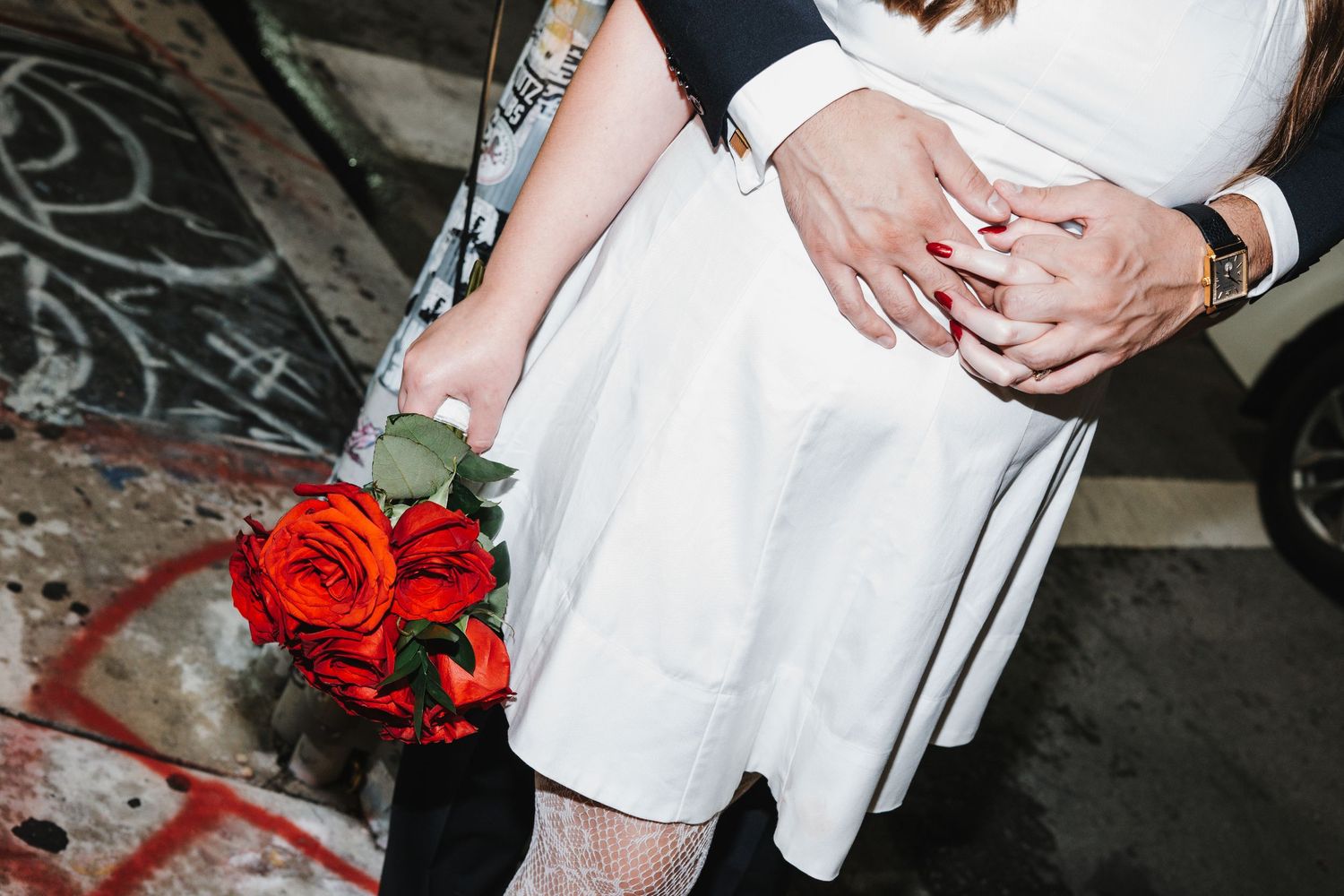 A couple holds hands during their wedding ceremony in front of red floral arrangements in a romantic setting.