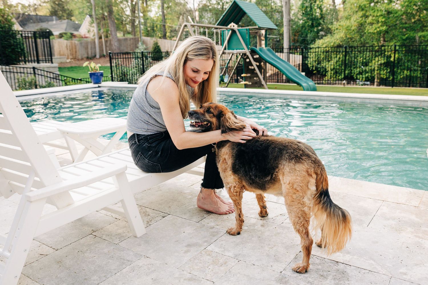 A German Shepherd mix dog being petted next to a backyard swimming pool with a playground set in the background.