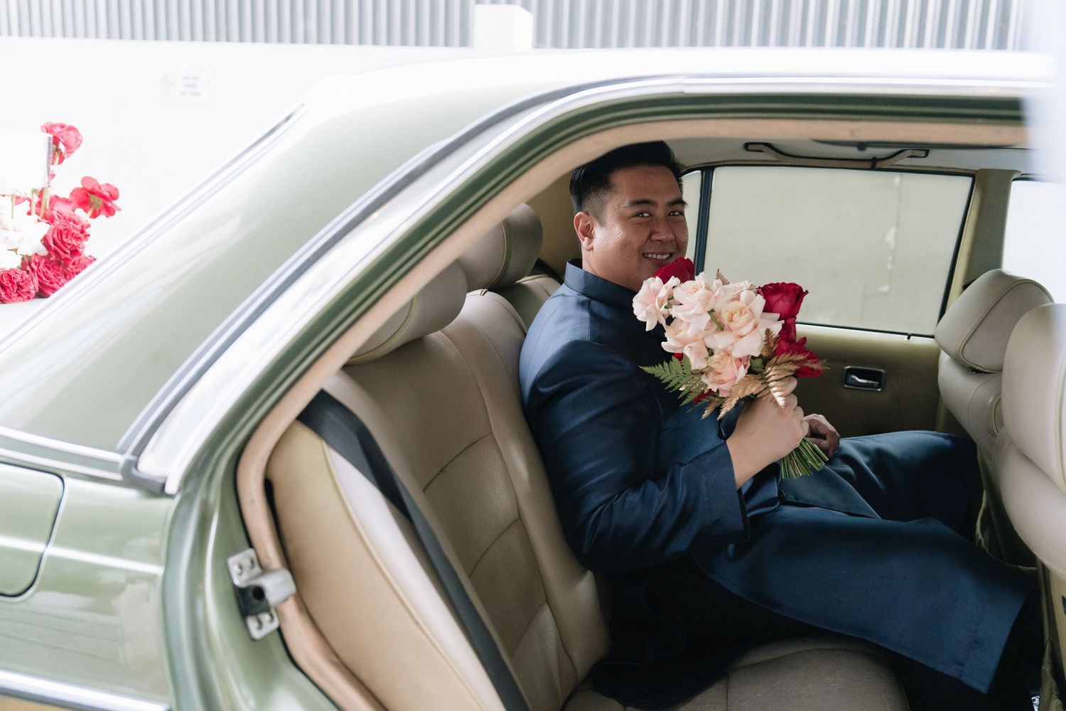 Groom in blue suit sitting in vintage car backseat holding pink and red bridal bouquet.