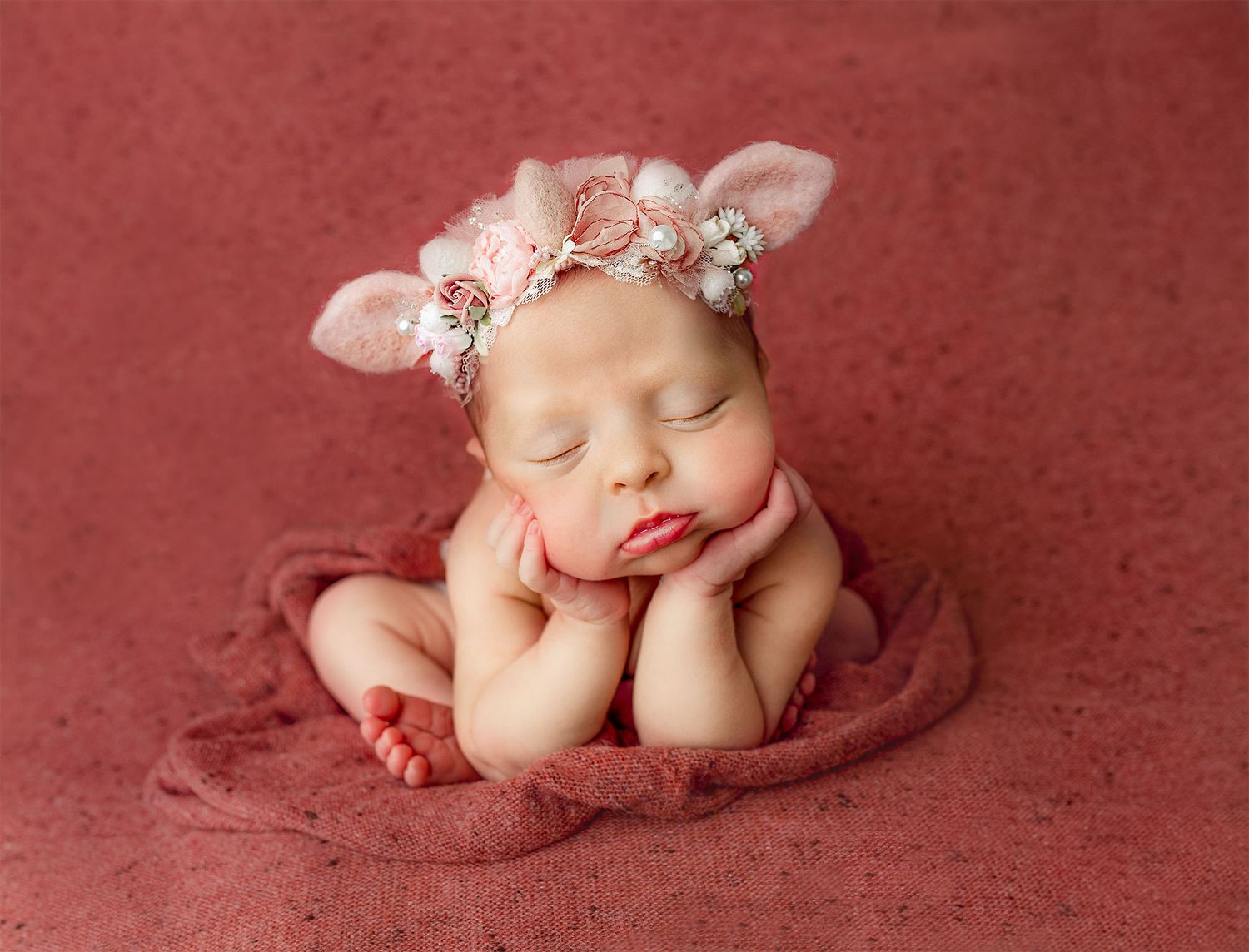 Newborn baby sleeping peacefully in fetal position on soft red backdrop with delicate floral headband.