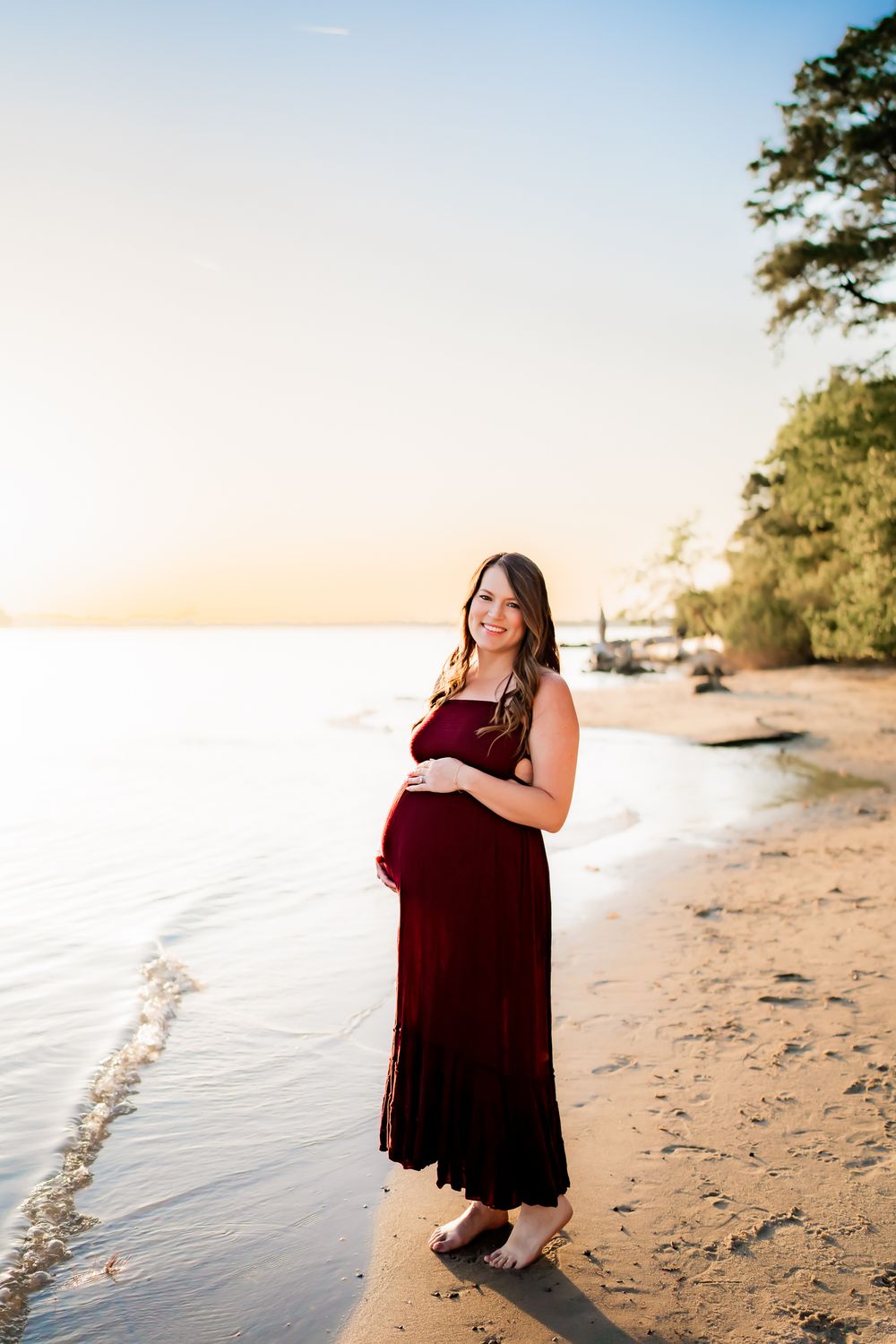 A young woman in a burgundy dress stands on a sandy beach at sunset, creating a serene coastal portrait.