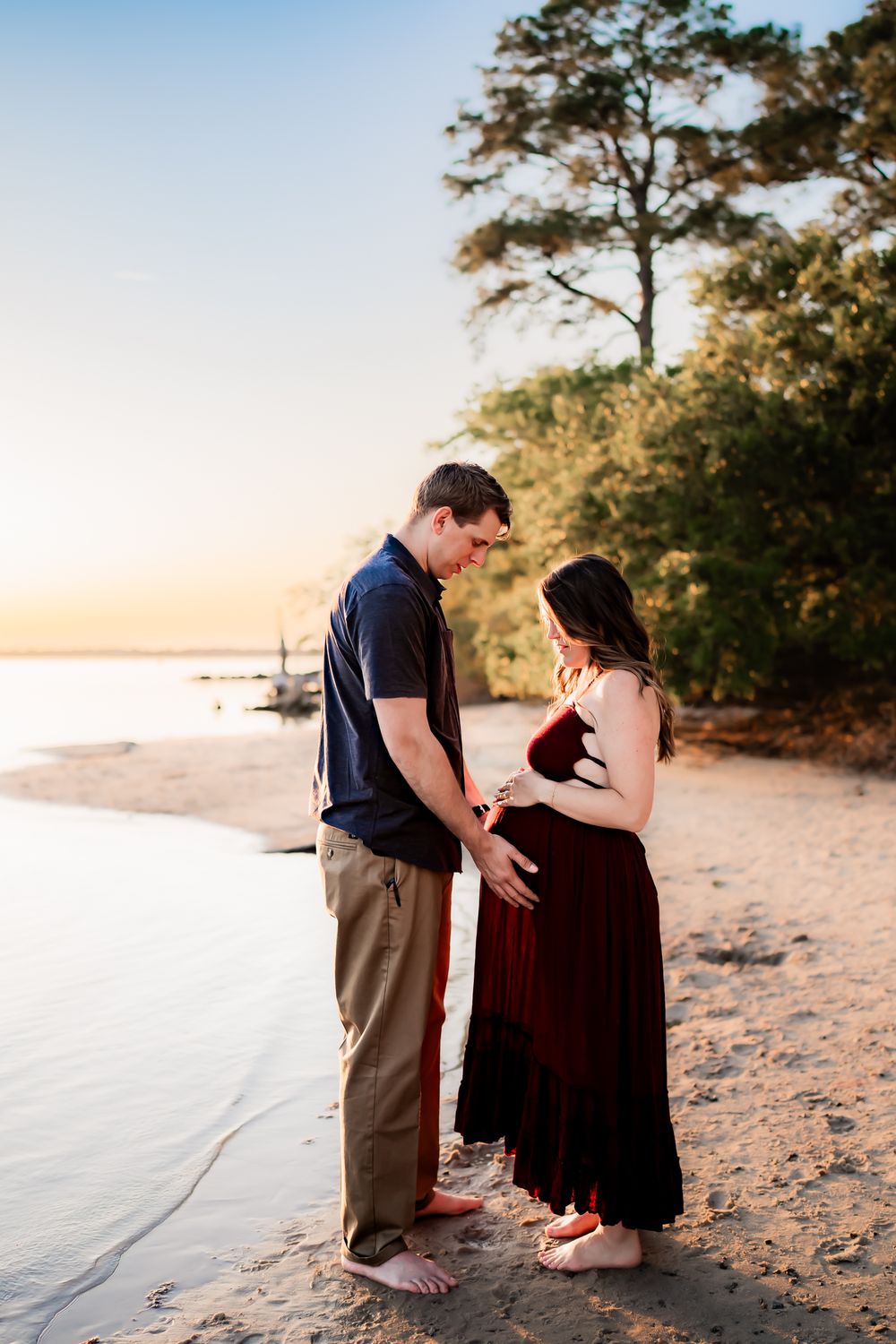 A couple shares a romantic moment during sunset on a sandy beach with pine trees in the background.