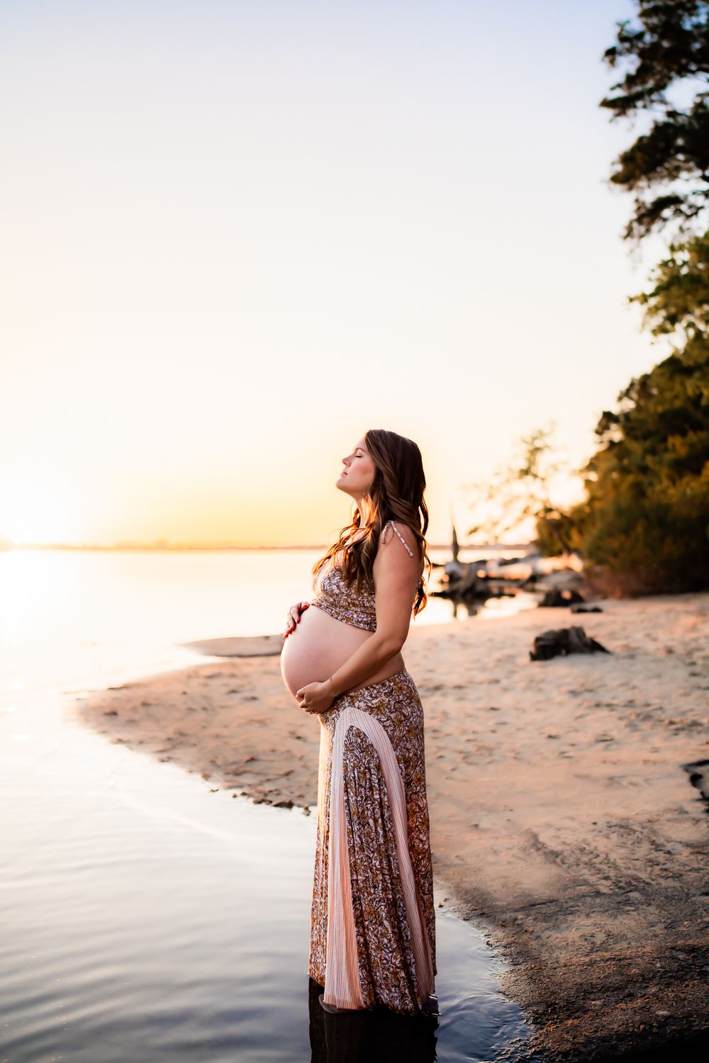 A maternity photo shoot on a sandy beach at sunset captures a serene moment by the water's edge.