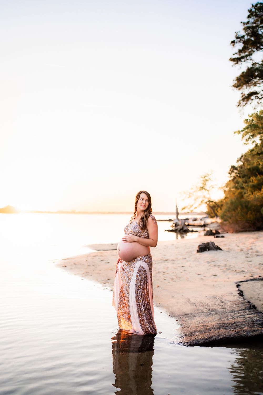 Expectant mother standing in shallow water at sunset wearing a floral dress during a beach photoshoot.