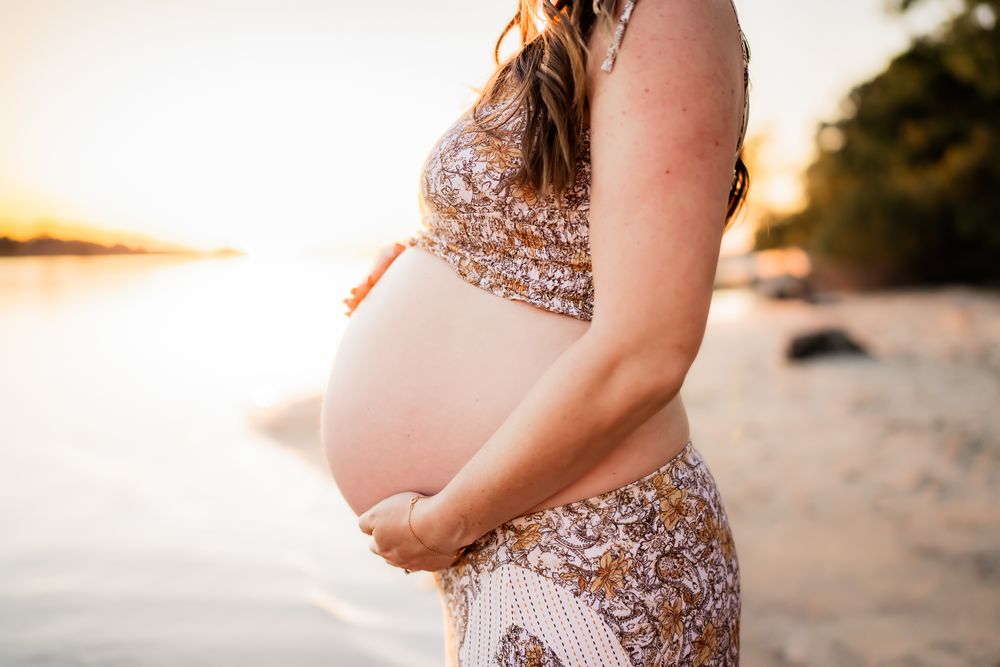 Silhouette of pregnant woman in floral dress holding belly at sunset on beach.