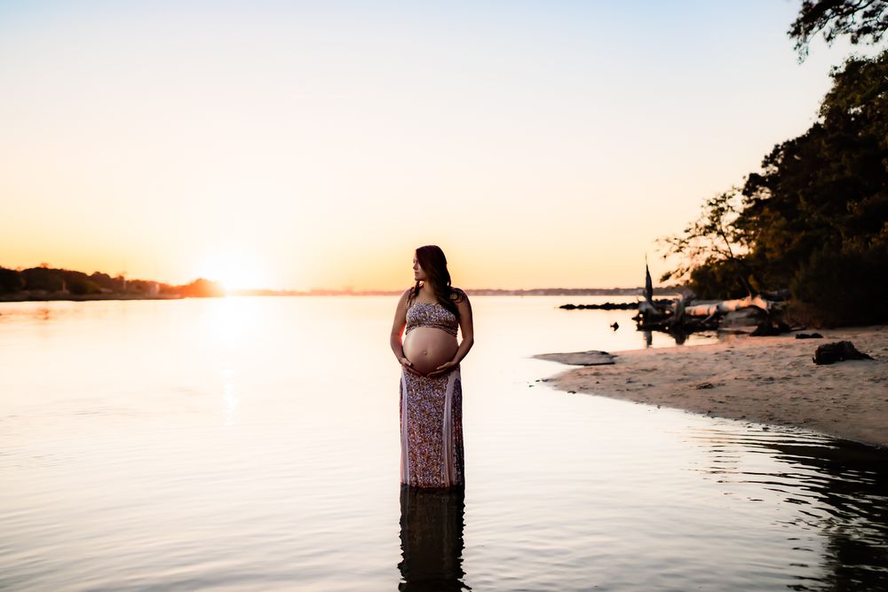 A couple stands on a beach at sunset with water and trees in the background creating a romantic silhouette.