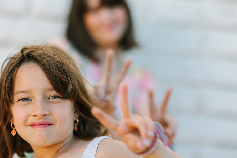 Two figures making peace signs and smiling at the camera against a light background.