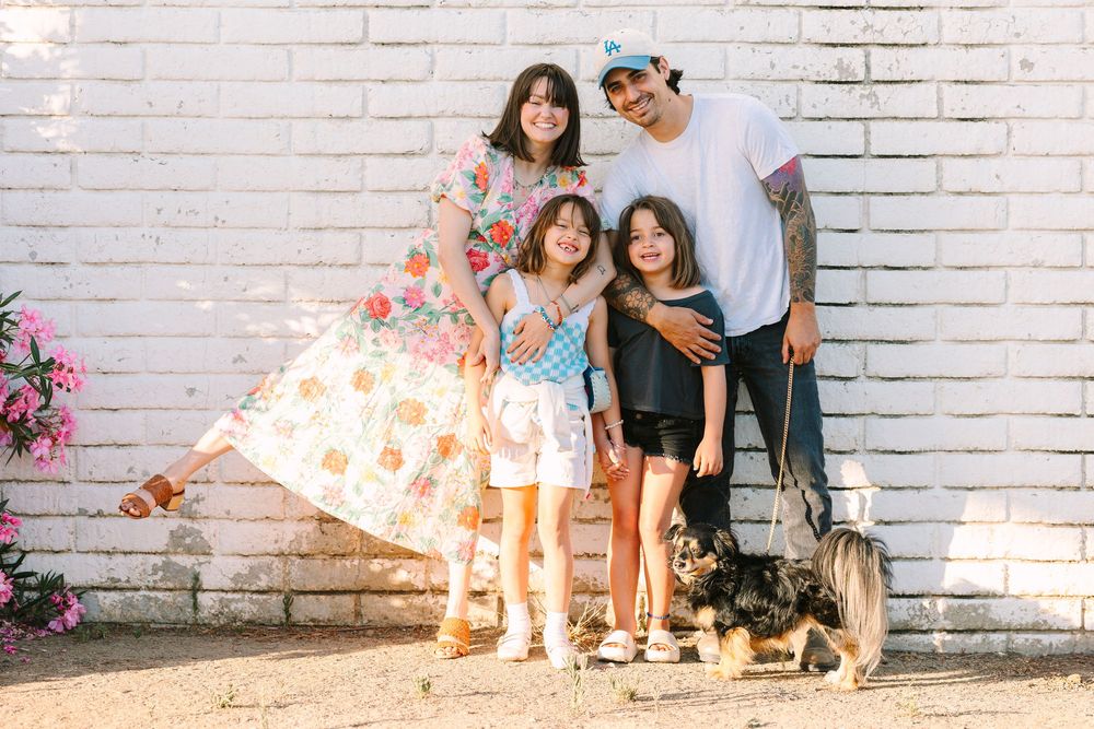 Family group poses together with their small dog against a white brick wall in sunlight.