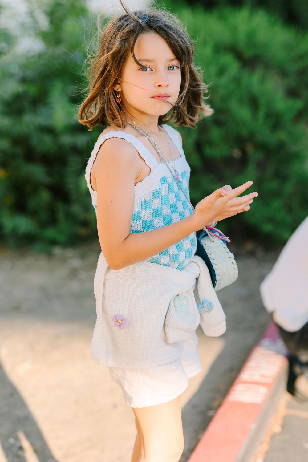 Person in blue checkered top with white bag poses against garden background.