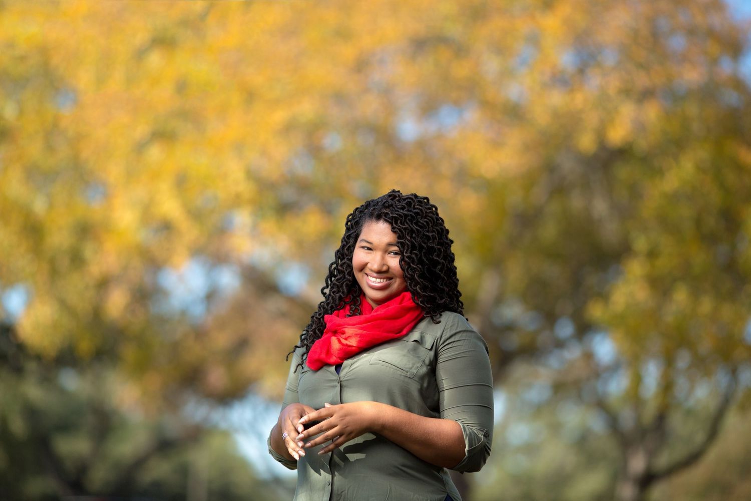A person wearing a red scarf and grey sweater smiles warmly against a backdrop of autumn foliage.