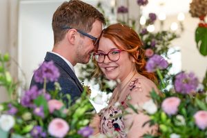 A romantic wedding couple embraces surrounded by pink and purple floral arrangements in a garden setting.
