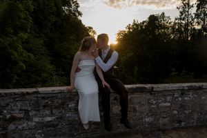 Bride and groom share a romantic sunset kiss on a stone wall surrounded by lush green trees at golden hour.
