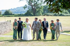 Wedding party members walk and laugh together in a line across a sunny grassy field on a summer day.