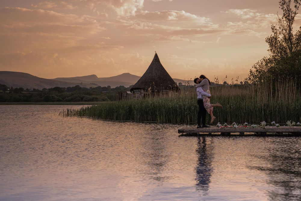 Couple embracing on a wooden dock at sunset beside a thatched hut, reeds, and a tranquil lake with mountain views.