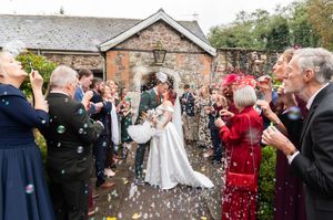 Wedding guests throw white confetti over a couple during an outdoor celebration at a stone building venue.