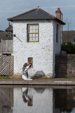 Bride and groom share a romantic dip kiss beside a white stone building reflected in calm canal waters.