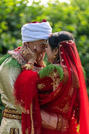 A romantic wedding moment captured between a couple in traditional red and cream attire against a lush green background.