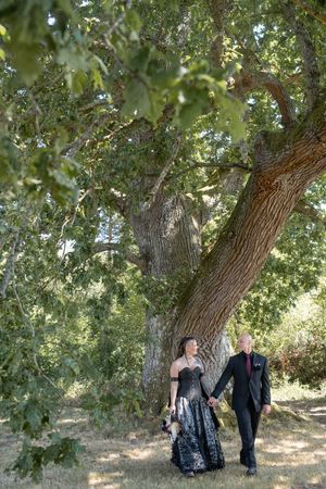 Couple in formal black attire holding hands under a large oak tree in a sunlit outdoor setting.