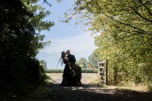 Couple sharing a romantic kiss silhouetted against a sunlit countryside path framed by lush green trees.