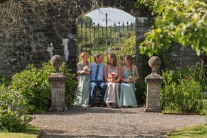 Wedding party sitting on a garden bench by an ornate iron gate, surrounded by lush greenery and stone pillars.