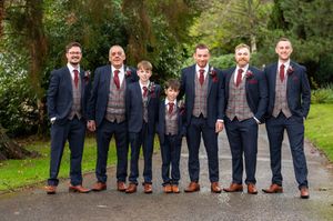 Wedding party members in navy suits show off their brown leather dress shoes while standing in a row on a garden path.