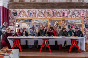 Medieval banquet reenactment with costumed guests seated at a long table before ornate heraldic tapestry.