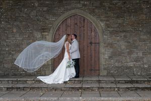 Bride and groom kissing before arched wooden church door, wedding veil flowing in wind, stone wall background.