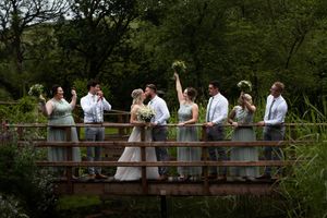 A wedding party group walks across a rustic wooden bridge surrounded by lush green foliage in a natural setting.
