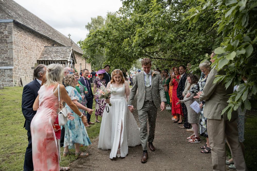 Bride and groom walk through a guest-lined path outside a stone church at a summer wedding celebration.