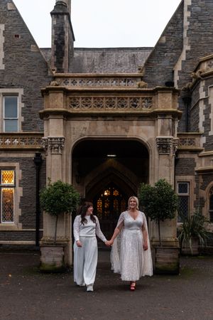 Two people in white dresses walk together outside a Gothic-style stone building with illuminated windows at dusk.