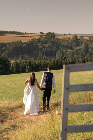 Bride and groom walking hand in hand through a sunlit green field with rolling countryside views.