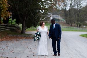 A couple in white and navy attire walking together along a tree-lined path in an autumn setting.