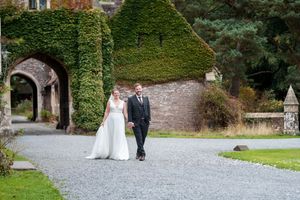 A wedding party walks down a stone path surrounded by lush gardens and trees at a historic venue.