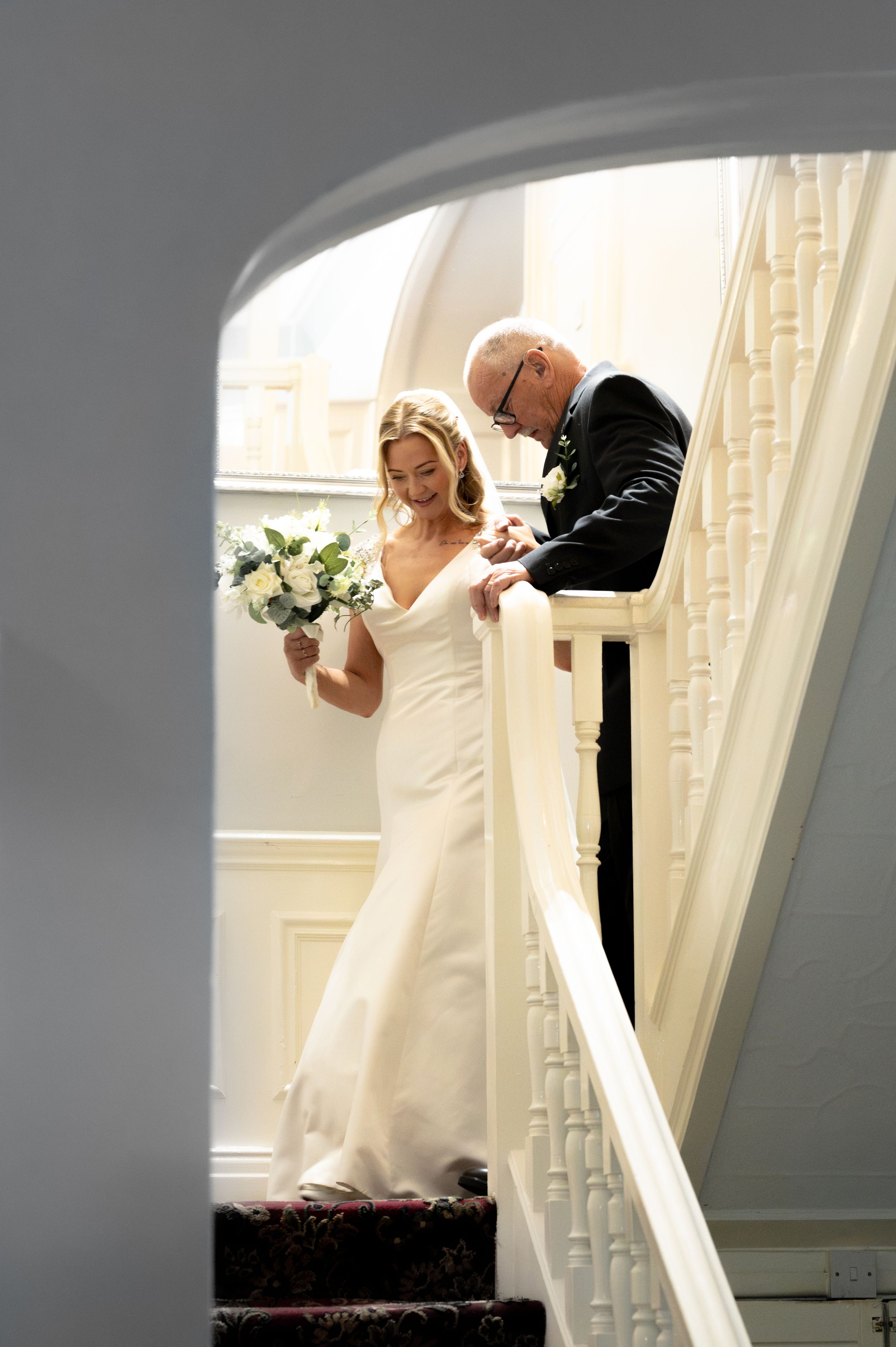 Bride in white gown holds bouquet beside suited man on elegant staircase during wedding preparations.