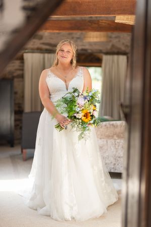 A bride in a white lace wedding gown holds a vibrant bouquet with sunflowers and greenery in a rustic indoor setting.