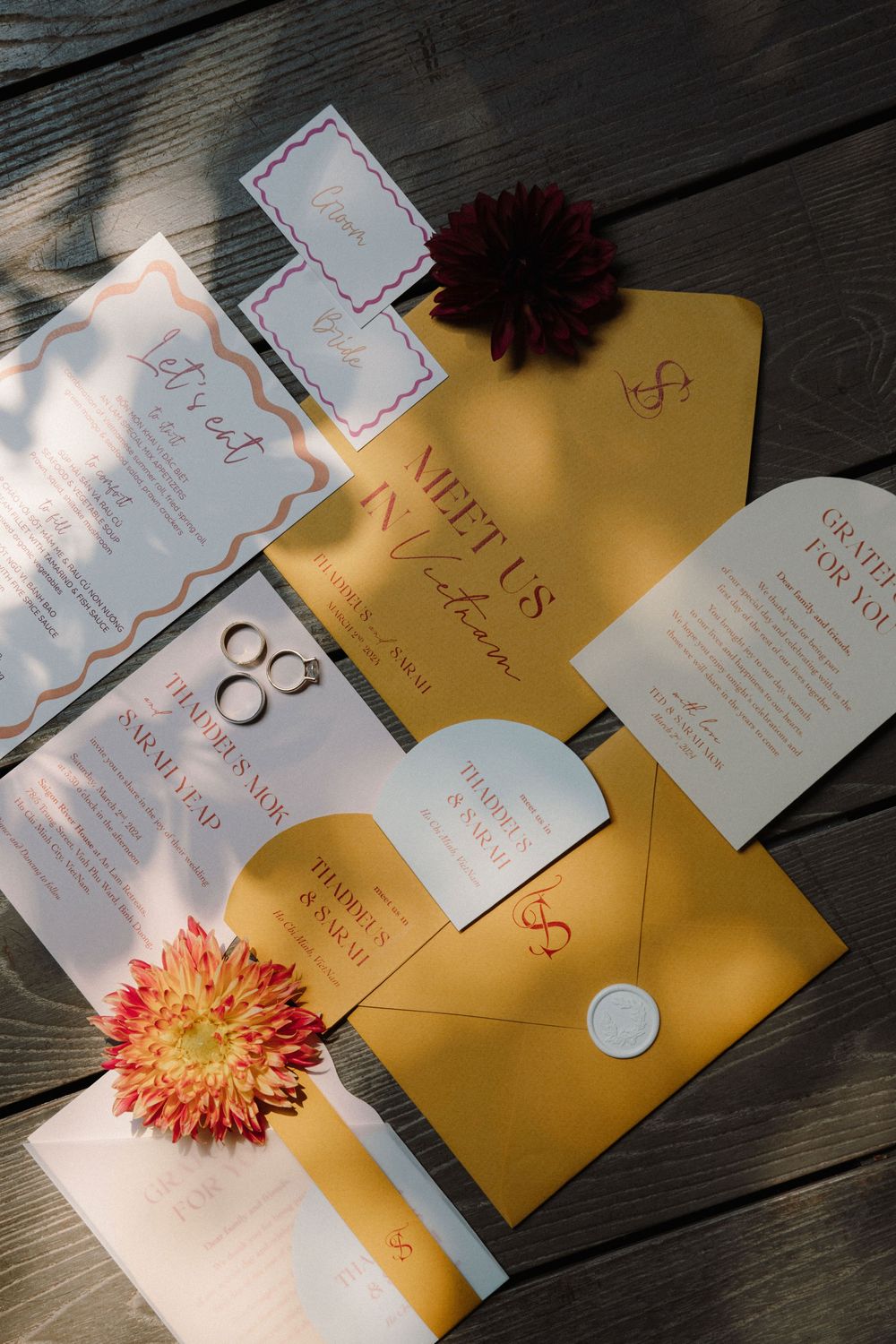Wedding invitations and envelopes arranged on dark wood with orange flowers and golden sunlight.