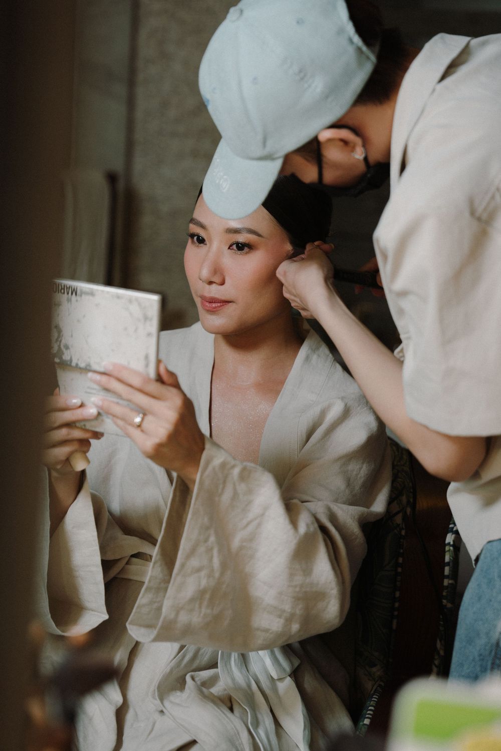 A person in a white robe holds a small cup while getting ready in what appears to be a dim, intimate setting.