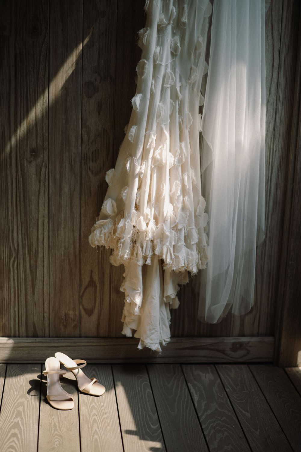 A flowing white wedding dress hanging in atmospheric lighting against a dark wooden wall.