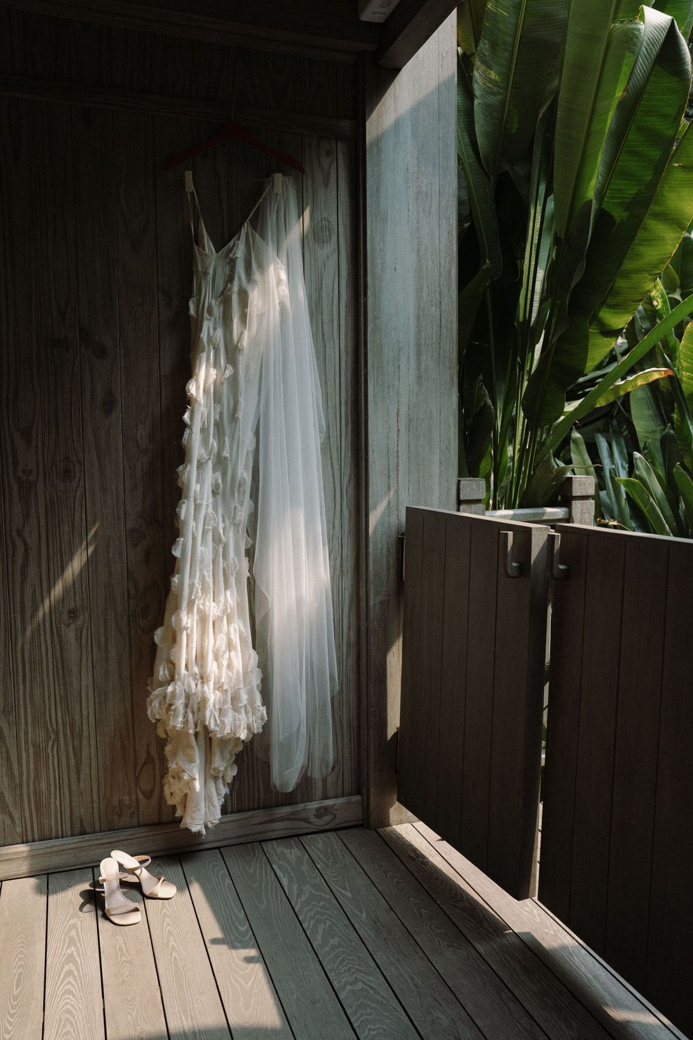White wedding dress photographed in natural light against wooden panels with tropical foliage.