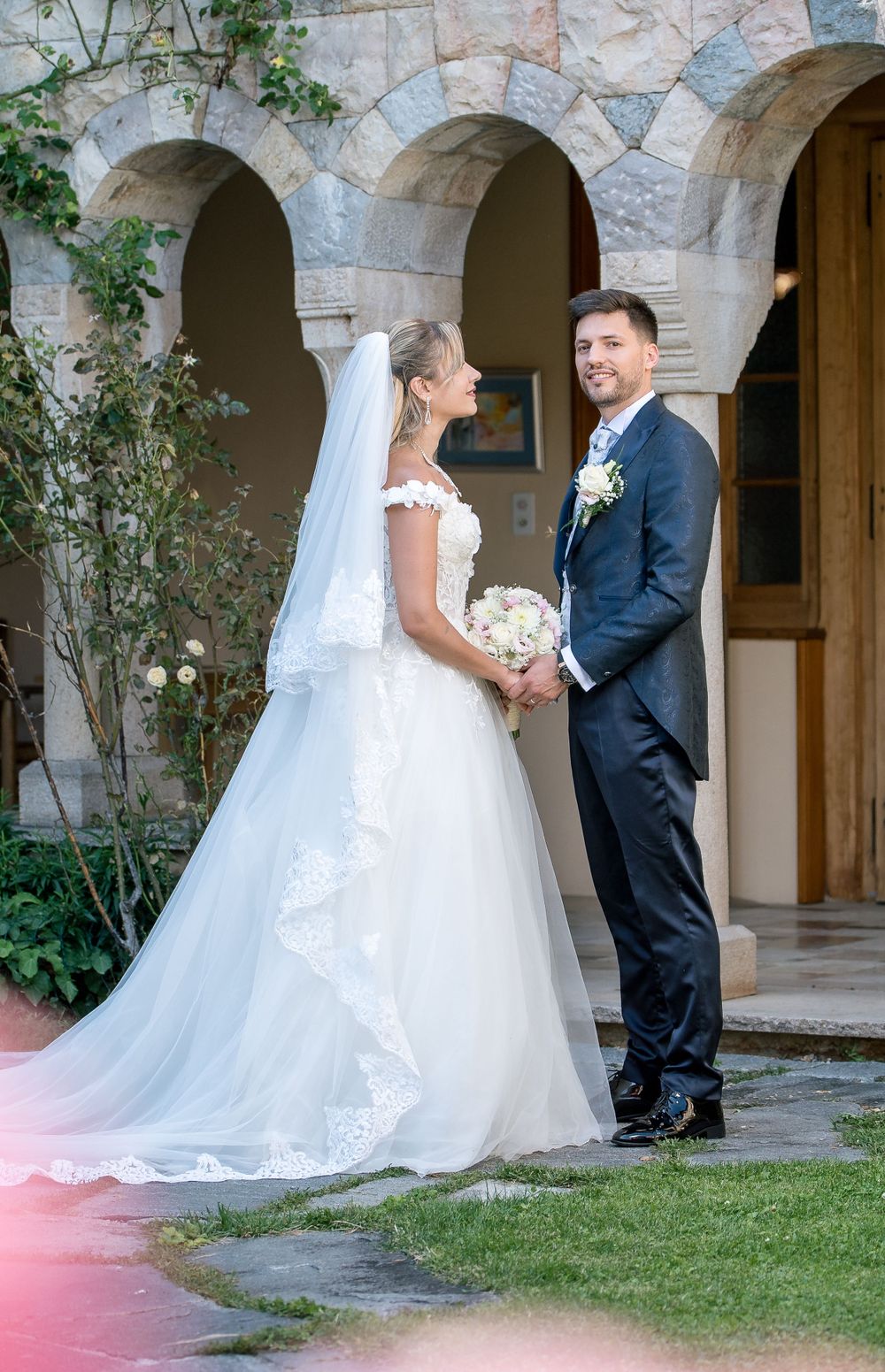 Newlyweds share romantic moment under stone arches with climbing vines.