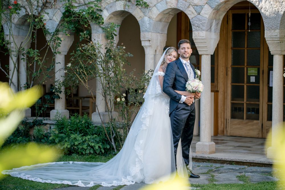Wedding couple embracing in stone courtyard with lush garden backdrop.